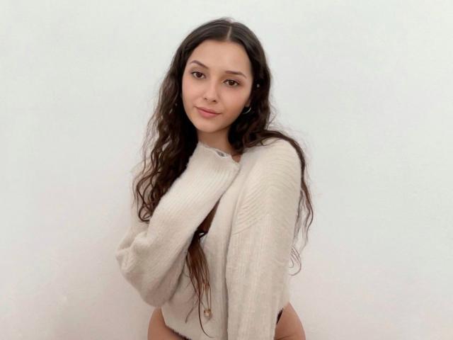 AngelEmma69 with long, wavy brown hair, wearing a white, ribbed sweater, covering her small, perky tits with her arms, standing against a plain, white background.