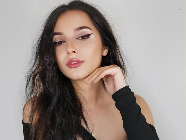 AngelEmma69, com cabelos pretos longos, delineador nos olhos, lábios rosados e uma blusa preta com um ombro à mostra, tocando o queixo com a mão. Ela tem um olhar sensual e sedutor..