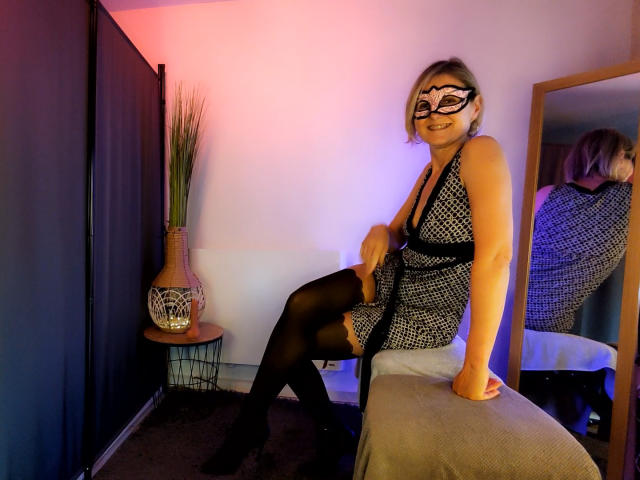 EroticaXEva con una máscara, vestido negro y medias, sentada en una silla, sonriendo, reflejo en el espejo, glúteos ligeramente visibles, piernas cruzadas, brazo sobre la silla, habitación con iluminación morada y rosa..