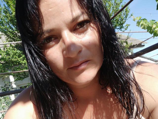 DeniseShiny, avec ses cheveux noirs mouillés, sa peau brun clair et ses yeux marrons, affichant une expression neutre, nue, baignée de soleil et brillante. L'arrière-plan comprend des arbres verts et des garde-corps en métal..