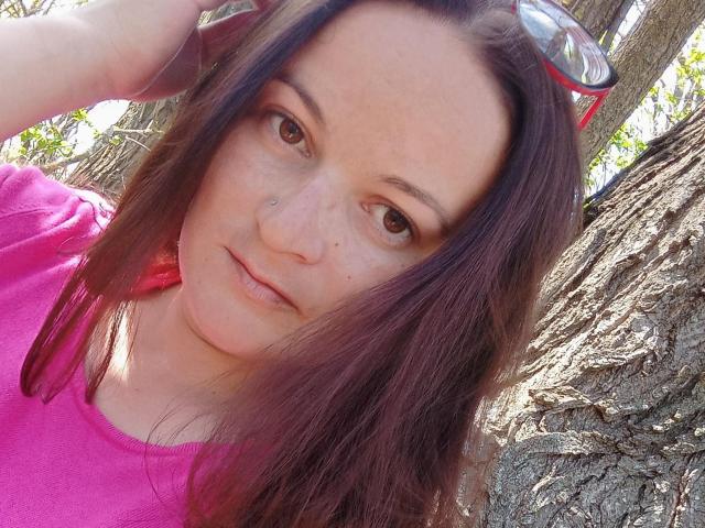 DeniseShiny with pink shirt, brown eyes, nose piercing, and red-rimmed sunglasses on head, outside in sunlight. Her purple-haired, slightly freckled face, small nose, and subtle smile.