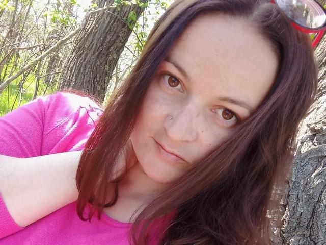DeniseShiny, con gli occhi marroni, un piercing al naso e una camicia rosa, ha i capelli castani lisci e un sorriso discreto, ed è appoggiata a un albero alla luce del sole. Il suo seno è parzialmente visibile..