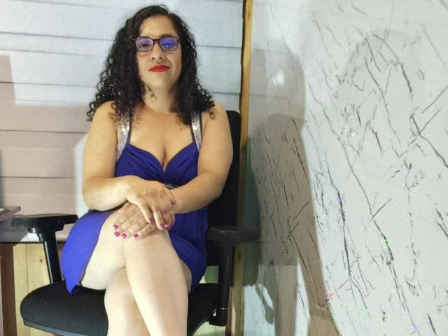 ShairaX with curly black hair, purple glasses, red lipstick, blue dress with silver straps, crossed legs, and pink nails, sitting on black chair, showing her tits.
