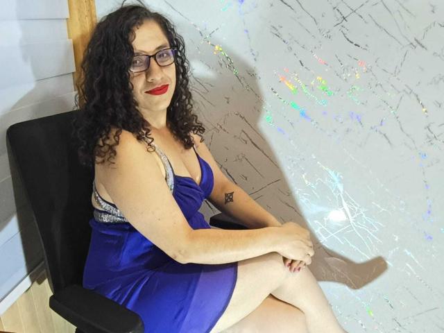 ShairaX with curly black hair, glasses, red lipstick, and blue dress, sitting on a black chair, showing her tits and a small tattoo on her right arm.