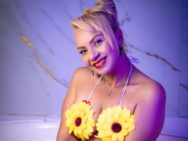 AnastasiaTaylor with blonde hair in a bun, smiling, wearing sunflower pasties covering her tits, red lipstick, and a white halter top, illuminated by purple and blue lights.