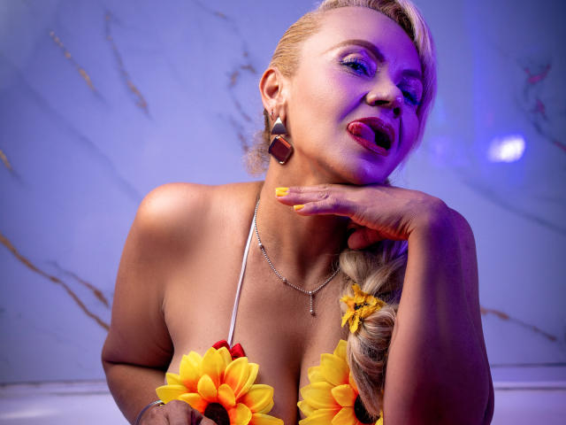AnastasiaTaylor with blonde hair in a braid, wearing flower bras, purple lights, and yellow nails, tongue out, hand on cheek, looking seductive, wearing geometric earrings, and a necklace.