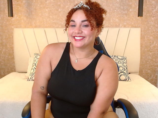 YeraCarey, con i capelli rossi ricci, una canottiera nera, che sorride, indossa una tiara, orecchini a cerchio, un piccolo tatuaggio a forma di cuore sul braccio sinistro, seduta su una sedia a rotelle, su un letto bianco con cuscini a motivi..