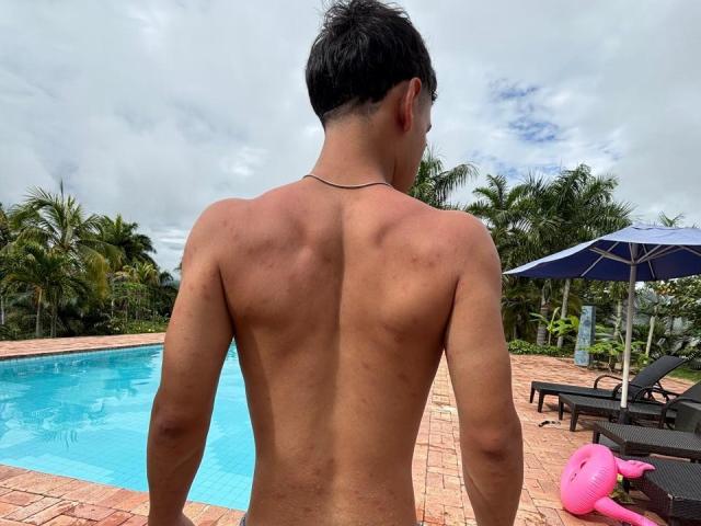 BrianOconor, ohne Hemd, gebräunte Haut, kurze schwarze Haare, schlank, kleines, dunkles Tattoo am Hals, steht neben einem blauen Pool, einem rosa Flamingo-Schwimmpflege, einem Sonnenschirm, Palmen, bewölkter Himmel..