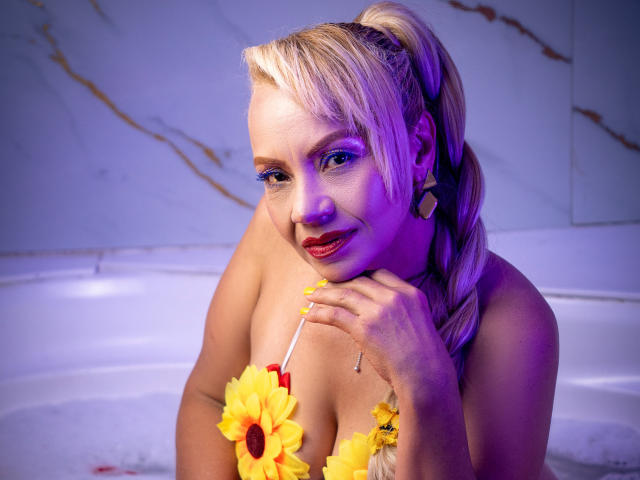 Anastasia Taylor, con la coda di cavallo bionda, fiori gialli sul décolleté, illuminazione viola, che tiene un girasole, unghie gialle, rossetto rosso, sorridente, con orecchini, in una vasca da bagno bianca, che guarda la fotocamera..