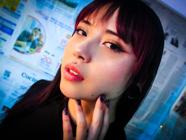 NaomyWallase with dark purple hair, pink lipstick, nose piercing, and glittery nails, looking at camera with hand on neck, blue and purple light, and soft focus.