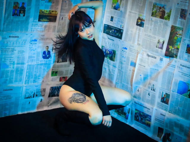 NaomyWallase, kneeling, wearing black long-sleeve bodysuit, showing tattoo on right thigh, dark hair, blue eyes, black boots, newspaper background, looking at camera, hand in hair.