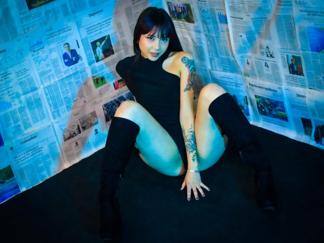 NaomyWallase with black turtleneck, black knee boots, and tattoos, sitting with legs spread, hands on floor, showing pussy, in a blue-lit room with newspaper background.