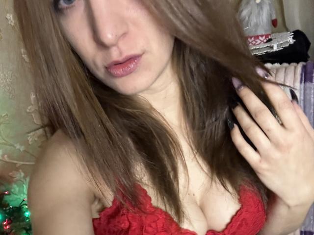 Cometomee, com cabelo castanho claro, lábios rosados, olhos azuis, e um sutiã de renda vermelho, exibindo seus seios, segurando o cabelo e olhando para a câmera..