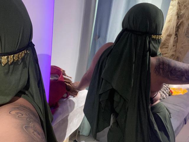 MiaHadid in a black burka with gold trim, showing a tattooed arm, bending over in front of a mirror, exposing her bare ass and back. Reflection shows her face.