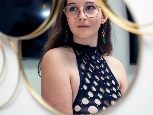 CoralieLaCoquine with glasses, wearing a black fishnet top revealing her tits, green earrings, and brown hair, reflected in a gold-framed mirror. She's smiling slightly.