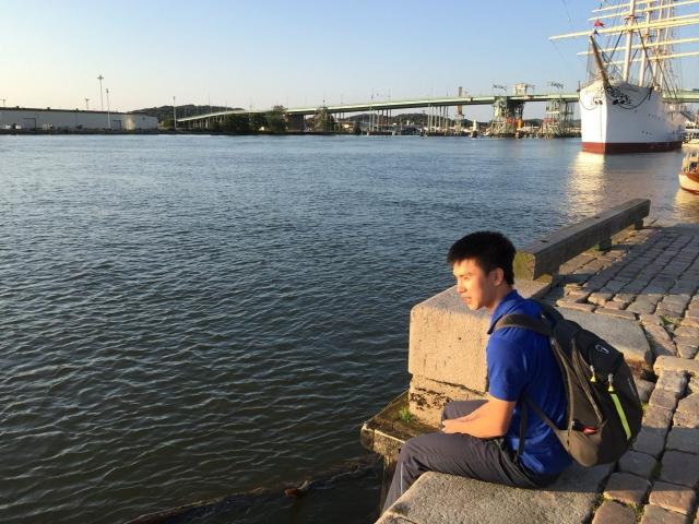 AngelWithAStarlight, sitting on a stone ledge by the water, wearing a blue shirt and grey backpack, looking back with short black hair, in a sunny, calm setting.