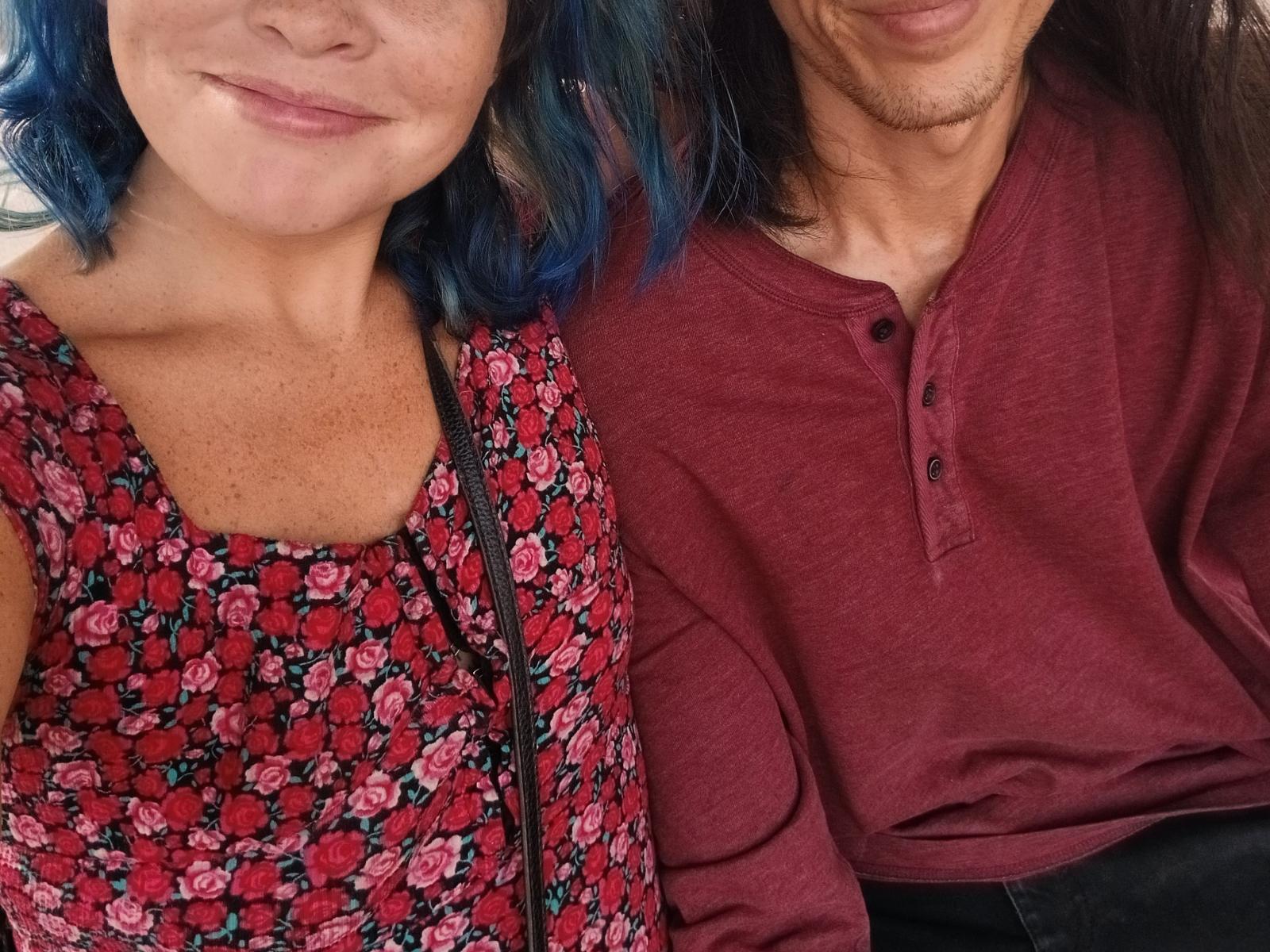 Due persone: una con capelli blu, lentiggini, che indossa un abito rosso con fiori, e un'altra con capelli castani, che indossa una maglia a maniche lunghe color borgogna, in un selfie. Entrambe hanno un sorriso leggero, e si vedono solo le loro parti superiori del corpo..