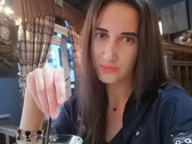 ModestMonika-hot with long brown hair, wearing a dark shirt, holding a spoon in her right hand, has a subtle smile, and a black necklace, in a dimly lit room.