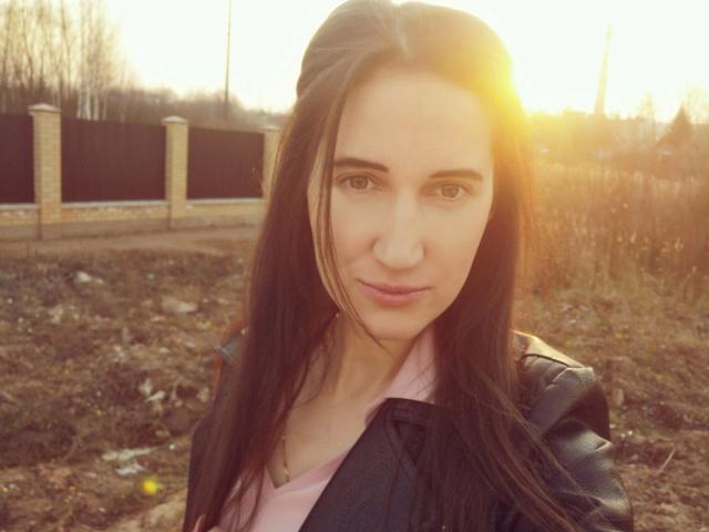 ModestMonika – attraktiv mit langen, braunen Haaren, trägt eine schwarze Lederjacke über einem pinken Hemd, Sonnenlicht im Hintergrund, ernster Gesichtsausdruck, weiches Licht, Schmutz und Gras im Vordergrund..