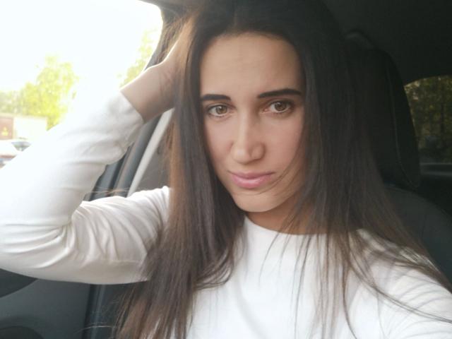 ModestMonika-hot with long brown hair, wearing a white long-sleeve shirt, raising her right hand, sitting in a car, looking at the camera with a slight smile.