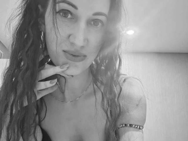 Black-and-white selfie of ModestMonika-hot with wavy hair, touching her chin, wearing a thin necklace and arm bracelet, showing her tits and subtle smile.