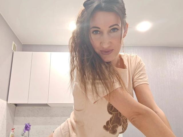 ModestMonika-hot with long brown hair, wearing a beige shirt, has blue eyes and is making a playful face. Her tits are visible under the shirt. Her arm is raised.