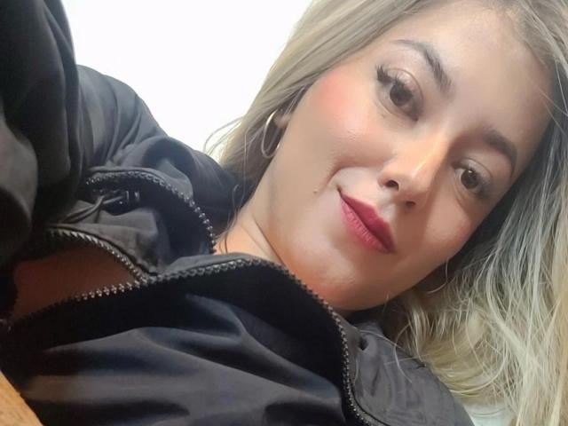KristenSexy has blonde hair, light skin, red lipstick, and is smiling in a black jacket, wearing hoop earrings, with a slight blush on her cheeks.