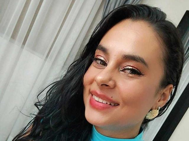 LunaSoler has long black hair, brown eyes with gold eyeliner, pink lipstick, a nose piercing, and is smiling with blue top. She's wearing gold earrings.
