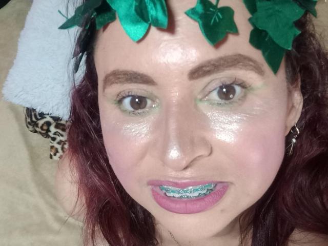 SherahaJade tiene una diadema con forma de hoja verde, lápiz labial rosa con brillo, sombra de ojos azul verdosa, ojos marrones y cabello rizado rojo. Su rostro brillante muestra una ligera sonrisa..