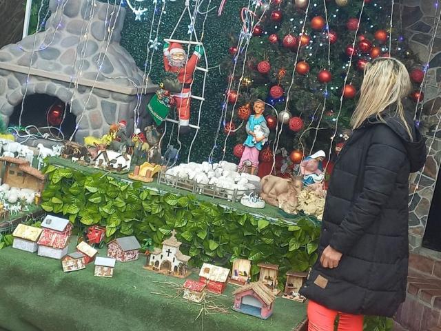 SherahaJade, con cabello rubio, chaqueta negra y abultada, pantalones naranja, de pie, mirando una exhibición navideña con muñecos de Santa Claus, adornos rojos, bolas blancas y pequeñas casas, sobre una mesa cubierta de hojas verdes..