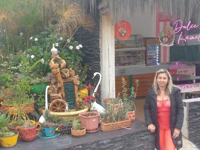 SherahaJade, con un busto de tamaño medio, está de pie sonriendo frente a una encimera de piedra negra, vestida con un vestido rojo y una chaqueta negra, rodeada de macetas de colores y una escultura de jardín caprichosa..