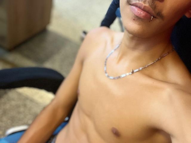 AndrewRock, shirtless with a silver chain, has a mustache, a lip piercing, and a toned, brown-skinned chest, sitting in a chair. Boobs: none, Ass: not visible, Tits: none.