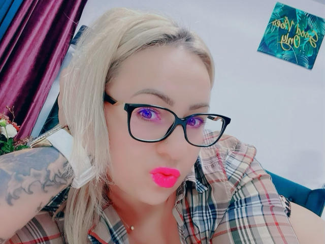 HotKrystall with blonde hair, purple eyes, black glasses, pink lipstick, plaid shirt, tattoo on arm, puckered lips, looking at camera. Purple curtains in background.