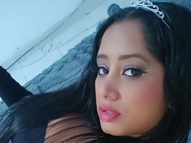 MariaCatalina with black hair, wearing a silver tiara, has pink lips, brown eyes with winged eyeliner, and a nose piercing, lying on a blue textured surface.