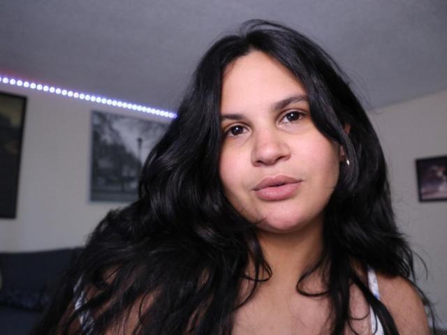 PrincessCata with long black wavy hair, medium brown skin, and pouty lips, wearing a white top, looking directly at the camera with a slight smile.