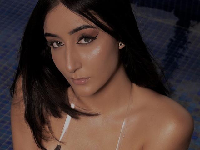 AlessiaRusso has shiny skin, dark brown hair, and wears a white top. Her eyeliner is thick, and she has a subtle smile. Her tits are slightly visible.