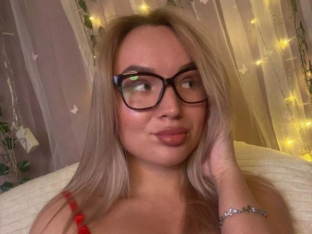 Jeanye-hot with blonde hair, glasses, and pouty lips, looking sideways, hand on cheek, wearing red beads, and a silver bracelet, in a softly lit room.