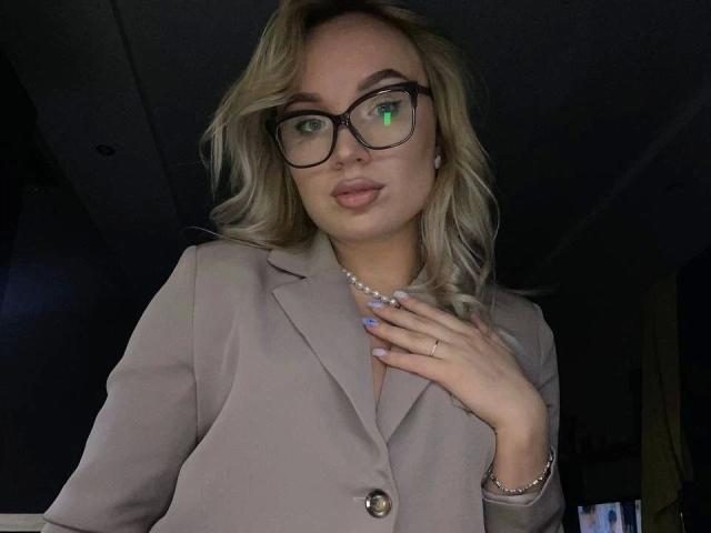 Jeanye-hot with blonde hair, glasses, and a pearl necklace, wearing a beige blazer, hand on chest, ring on finger, light purple nails, subtle makeup, green eye reflection.