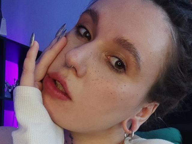 Eva Ragen tem um piercing no nariz, sardas e sobrancelhas escuras. Ela está tocando sua bochecha com unhas longas e metálicas, usando uma manga branca e tem piercings nas orelhas..