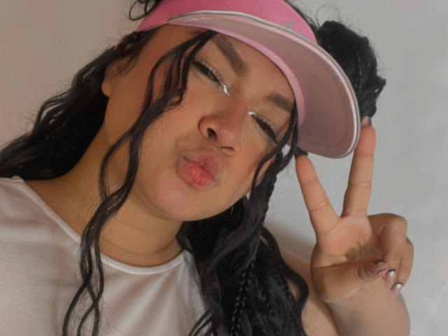 LillyMayer with black hair, pink visor, and white shirt, making a peace sign while puckering her lips, eyes winking. She's got a cute, sexy, and flirty look.