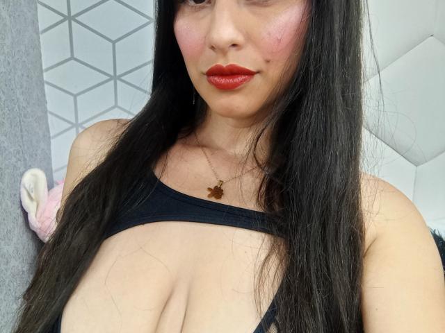MadissonFontaine with long black hair, red lipstick, and a black top showing her large, bare tits, wearing a small gold necklace, in a selfie.