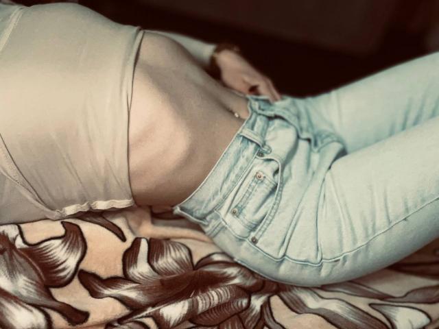 NikaEmbert's slim, pale, flat-ass in tight, light-blue jeans, wearing a beige, cut-out top, lying on a patterned bed. Visible belly button.