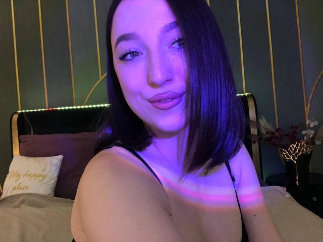 MonAmourR hat eine violett schimmernde Haut, mittelgroße Brüste und dunkelbraunes, schulterlanges Haar. Sie lächelt mit pinkfarbener Lippenstift und trägt ein schwarzes Top mit Spaghetti-Trägern, in einem schwach beleuchteten Schlafzimmer..