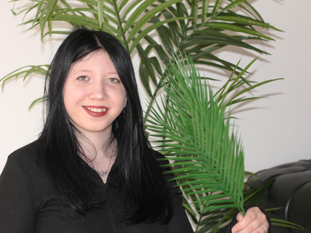 LolaBrook with black hair, fair skin, red lipstick, black top, holding green palm leaf, smiling, standing in front of black leather couch, white wall background.