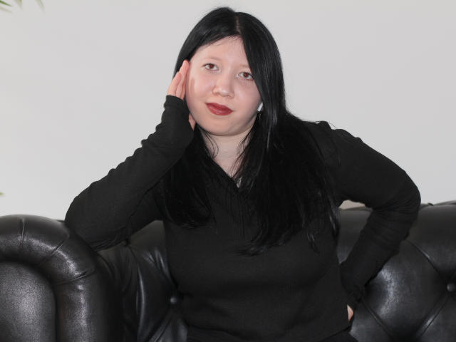LolaBrook with black hair, wearing a black long-sleeve shirt, leaning on a black leather couch, hand on cheek, red lipstick, white earbuds, and a relaxed, slightly sexy look.