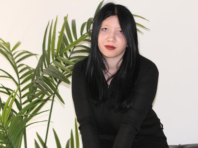 LolaBrook with black hair, wearing a black long-sleeve shirt, leaning forward, looking at camera, red lipstick, green potted plant to the left, white wall background.