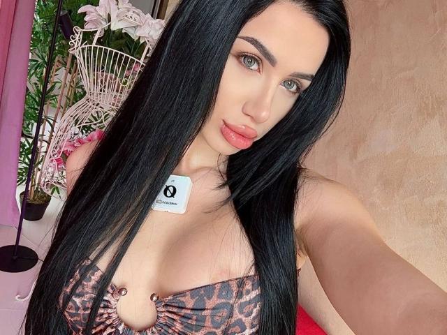 JennaDay has long black hair, green eyes, and pink lips. She's wearing a leopard print top with a keyhole cutout showing her tits. She's holding a white card.