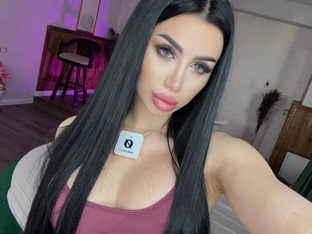 JennaDay has long black hair, green eyes, pink lipstick, and wears a pink tank top, showing her tits with a Quora sticker on her chest. She's taking a selfie.