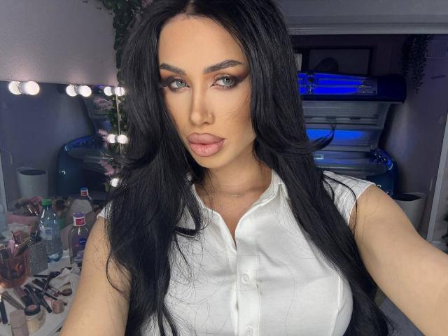 JennaDay with long black hair, full lips, and blue eyes, wearing a white button-up shirt, showing her tits, in a room with makeup and bottles on a table.