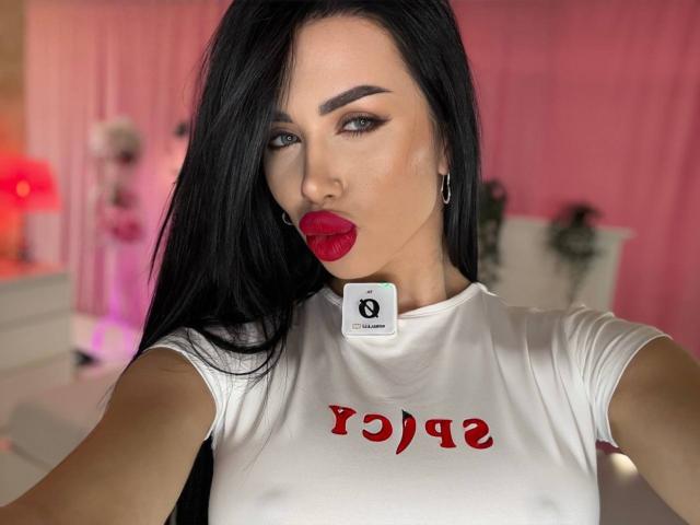 JennaDay with black hair, red lipstick, and white shirt with red text, showing a "Q" sticker on her neck, looking at the camera with a pouty expression.